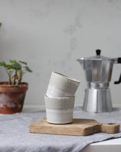 Stack of ceramic cups on a wooden cutting board with a metal coffee percolator in the background.